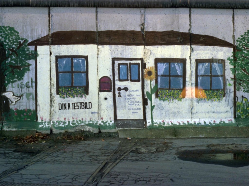 Ein über mehrere Mauersegmente gemaltes Bild zeigt ein weißes Haus zwischen zwei Bäumen. Das Haus hat eine Tür, drei Fenster und einen Briefkasten. Unter dem Fenster links steht: Din A Testbild. Neben der Tür ist eine Sonnenblume gemalt.