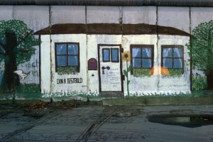 Ein &uuml;ber mehrere Mauersegmente gemaltes Bild zeigt ein wei&szlig;es Haus zwischen zwei B&auml;umen. Das Haus hat eine T&uuml;r, drei Fenster und einen Briefkasten. Unter dem Fenster links steht: Din A Testbild. Neben der T&uuml;r ist eine Sonnenblume gemalt.