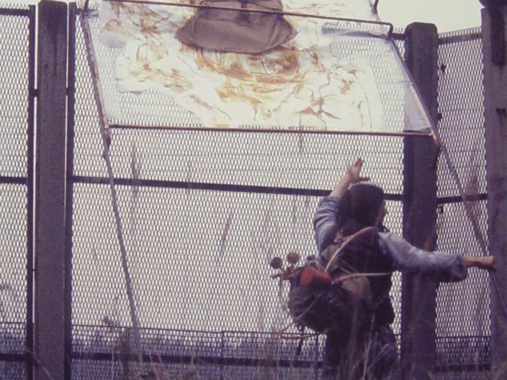 Stephan Elsner ist von Hinten aufgenommen. Er hält das Bild, das oben auf dem Zaun aufliegt mit einer Hand und greift mit der anderen danach.