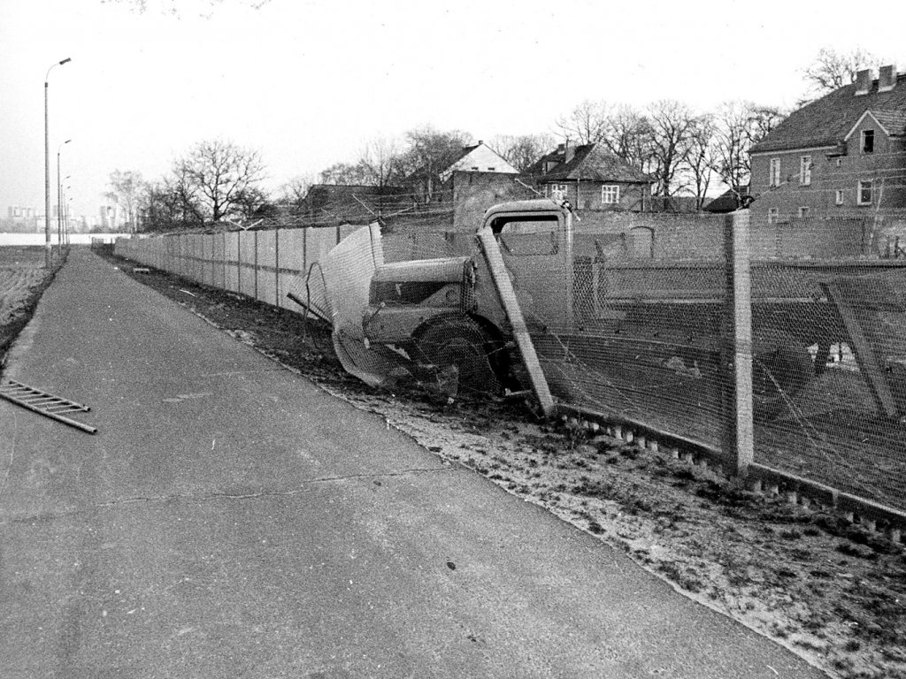 Im Bildzentrum sieht man einen LKW, der teilweise durch den Grenzzaun durchgebrochen ist. Auf dem Postenweg liegt noch die zurückgelassene Fluchtleiter.