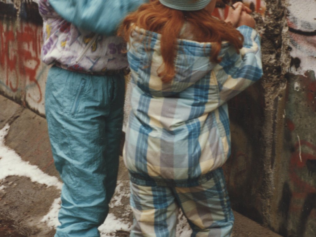 Zwei kleine Mädchen in bunter Winterkleidung bearbeiten die bereits teilweise zerstörte und mit Graffiti überzogene Berliner Mauer mit Werkzeugen.
