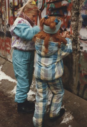 Zwei kleine Mädchen in bunter Winterkleidung bearbeiten die bereits teilweise zerstörte und mit Graffiti überzogene Berliner Mauer mit Werkzeugen.