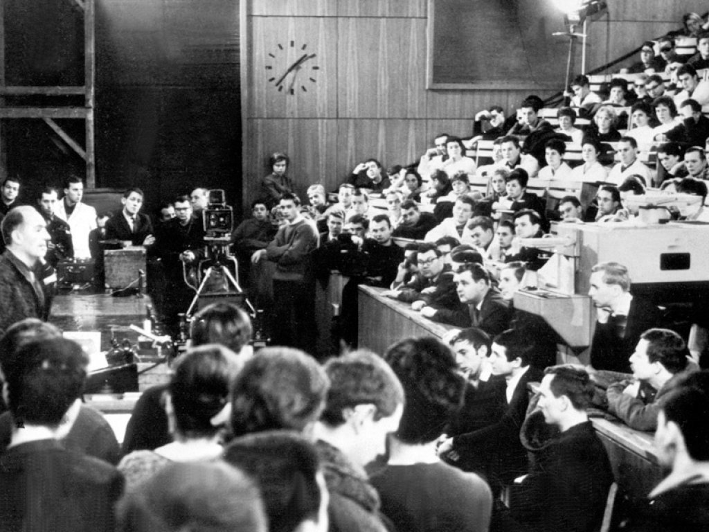 Robert Havemann bei seiner letzten Vorlesung in der Ost-Berliner Humboldt-Universität, 17. Januar 1964 Aufnahme eines vollen Hörsaals mit Havemann am linken Bildrand. Links und rechts neben dem Rednerpult drängen sich Zuhörerinnen und Zuhörer. Im Bildhintergrund ist eine Kamera aufgebaut.