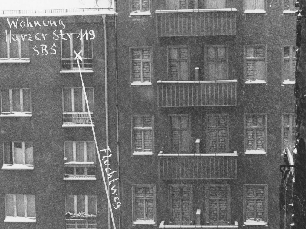 Frontalaufnahme von zwei aneinandergebauten Wohnhäusern. Die Fenster des rechten Hauses sind zugemauert. Am unteren Bildrand trennt eine Mauer die Fläche vor den Häusern. Neben einem Fenster im vierten Stock des linken Hauses ist handschriftlich notiert: Wohnung Harzer Str 119 SBS. Von hier führt ein Pfeil mit der Beschriftung: Fluchtweg schräg nach unten rechts, wo steht: Aufsprung auf Westberliner Gebiet.