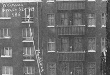 Frontalaufnahme von zwei aneinandergebauten Wohnhäusern. Die Fenster des rechten Hauses sind zugemauert. Am unteren Bildrand trennt eine Mauer die Fläche vor den Häusern. Neben einem Fenster im vierten Stock des linken Hauses ist handschriftlich notiert: Wohnung Harzer Str 119 SBS. Von hier führt ein Pfeil mit der Beschriftung: Fluchtweg schräg nach unten rechts, wo steht: Aufsprung auf Westberliner Gebiet.