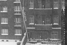 Frontalaufnahme von zwei aneinandergebauten Wohnhäusern. Die Fenster des rechten Hauses sind zugemauert. Am unteren Bildrand trennt eine Mauer die Fläche vor den Häusern. Neben einem Fenster im vierten Stock des linken Hauses ist handschriftlich notiert: Wohnung Harzer Str 119 SBS. Von hier führt ein Pfeil mit der Beschriftung: Fluchtweg schräg nach unten rechts, wo steht: Aufsprung auf Westberliner Gebiet.