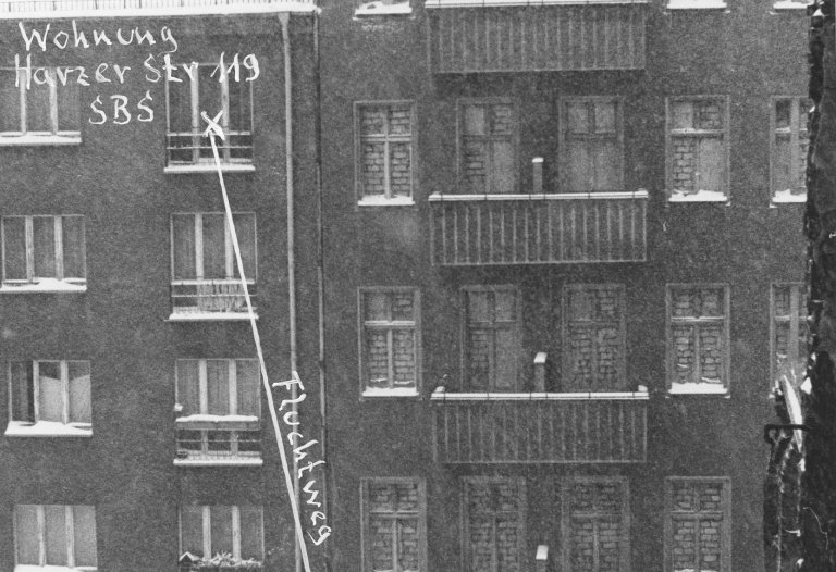 Frontalaufnahme von zwei aneinandergebauten Wohnhäusern. Die Fenster des rechten Hauses sind zugemauert. Am unteren Bildrand trennt eine Mauer die Fläche vor den Häusern. Neben einem Fenster im vierten Stock des linken Hauses ist handschriftlich notiert: Wohnung Harzer Str 119 SBS. Von hier führt ein Pfeil mit der Beschriftung: Fluchtweg schräg nach unten rechts, wo steht: Aufsprung auf Westberliner Gebiet.