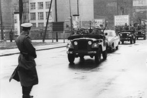 Links im Vordergrund steht ein Uniformierter mit dem Rücken zur Kamera. Von rechts kommend fährt eine Fahrzeugkolonne durch die Grenzsperren im Hintergrund auf die Bildmitte zu. Das zweite Auto ist ein Zivilwagen, vor und hinter ihm fahren Soldaten in Jeeps.