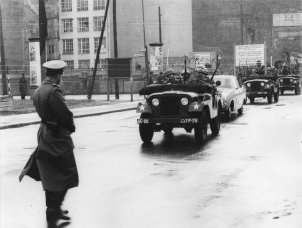 Links im Vordergrund steht ein Uniformierter mit dem Rücken zur Kamera. Von rechts kommend fährt eine Fahrzeugkolonne durch die Grenzsperren im Hintergrund auf die Bildmitte zu. Das zweite Auto ist ein Zivilwagen, vor und hinter ihm fahren Soldaten in Jeeps.