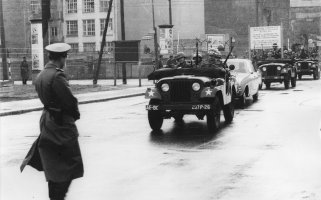 Links im Vordergrund steht ein Uniformierter mit dem Rücken zur Kamera. Von rechts kommend fährt eine Fahrzeugkolonne durch die Grenzsperren im Hintergrund auf die Bildmitte zu. Das zweite Auto ist ein Zivilwagen, vor und hinter ihm fahren Soldaten in Jeeps.
