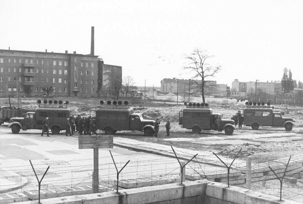 Im Vordergrund verläuft die Mauer aus Hohlblocksteinen und Stacheldraht, dahinter stehen vier Lastwagen mit Lautsprechern auf den Dächern. Einige Soldaten stehen daneben.