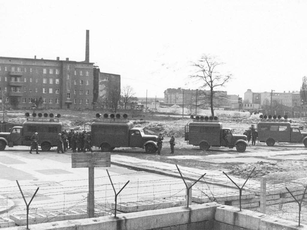 Im Vordergrund verläuft die Mauer aus Hohlblocksteinen und Stacheldraht, dahinter stehen vier Lastwagen mit Lautsprechern auf den Dächern. Einige Soldaten stehen daneben.