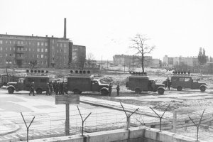 Im Vordergrund verläuft die Mauer aus Hohlblocksteinen und Stacheldraht, dahinter stehen vier Lastwagen mit Lautsprechern auf den Dächern. Einige Soldaten stehen daneben.