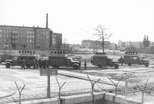 Im Vordergrund verläuft die Mauer aus Hohlblocksteinen und Stacheldraht, dahinter stehen vier Lastwagen mit Lautsprechern auf den Dächern. Einige Soldaten stehen daneben.