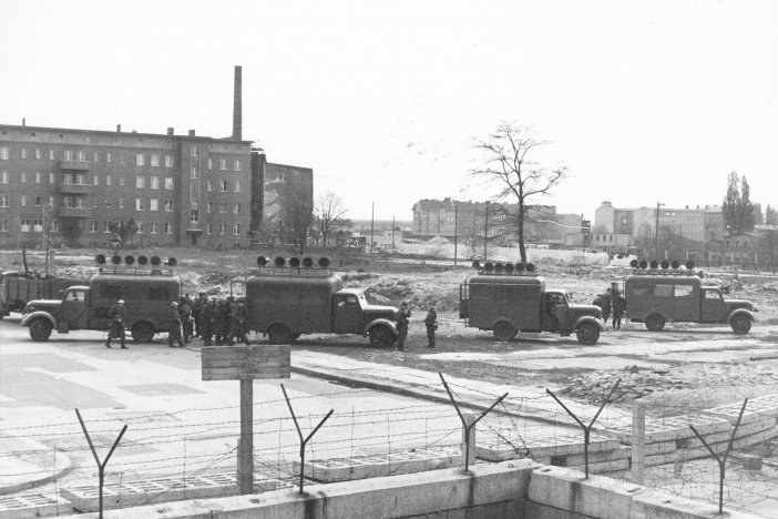 Im Vordergrund verläuft die Mauer aus Hohlblocksteinen und Stacheldraht, dahinter stehen vier Lastwagen mit Lautsprechern auf den Dächern. Einige Soldaten stehen daneben.