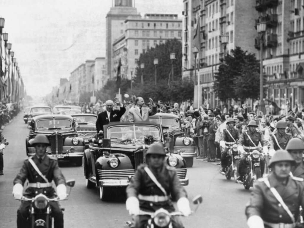 Freundschaftsbesuche von Nikita Chruschtschow anlässlich des 70. Geburtstages von Walter Ulbricht (hier Karl-Marx-Allee); Aufnahme 28. Juni 1963 Polizisten auf Motorrädern fahren auf die Kamera zu. Hinter ihnen fährt ein Auto mit offenem Verdeck, in dem Chruschtschow und Ulbricht stehen und winken. Dahinter folgen weitere Autos. Die Straße ist von winkenden Menschen gesäumt.