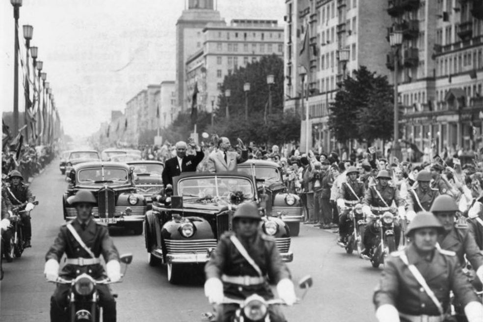 Polizisten auf Motorr&auml;dern fahren auf die Kamera zu. Hinter ihnen f&auml;hrt ein Auto mit offenem Verdeck, in dem Chruschtschow und Ulbricht stehen und winken. Dahinter folgen weitere Autos. Die Stra&szlig;e ist von winkenden Menschen ges&auml;umt.
