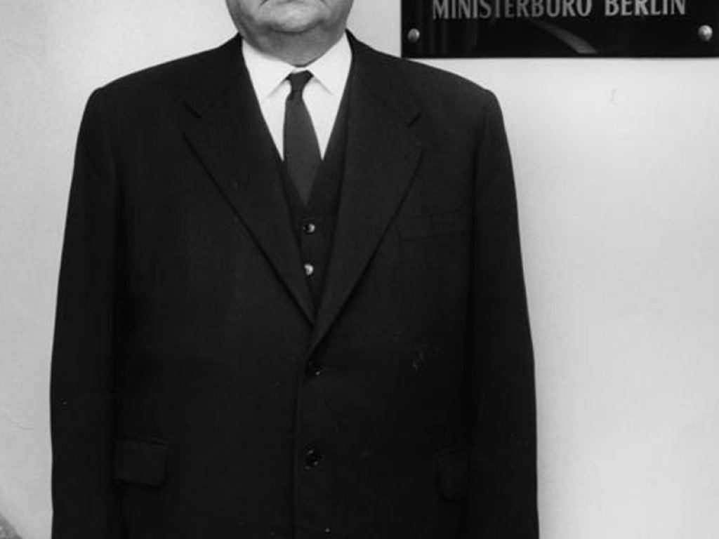 Ernst Lemmer steht in einem Treppenhaus und blickt direkt und ernst in die Kamera. Hinter ihm an der Wand steht auf einem Schild: Der Bundesminister für gesamtdeutsche Fragen. Ministerbüro Berlin.
