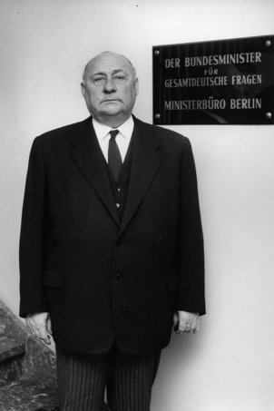 Ernst Lemmer steht in einem Treppenhaus und blickt direkt und ernst in die Kamera. Hinter ihm an der Wand steht auf einem Schild: Der Bundesminister für gesamtdeutsche Fragen. Ministerbüro Berlin.