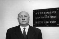 Ernst Lemmer steht in einem Treppenhaus und blickt direkt und ernst in die Kamera. Hinter ihm an der Wand steht auf einem Schild: Der Bundesminister für gesamtdeutsche Fragen. Ministerbüro Berlin.