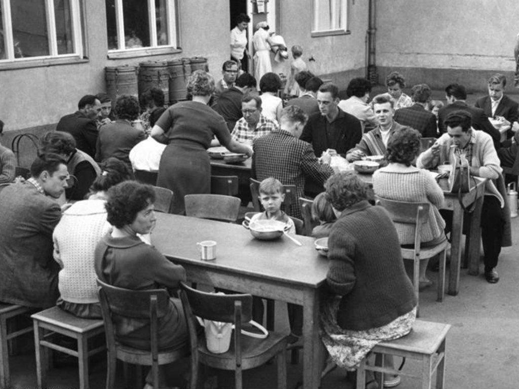 Das Notaufnahmelager Marienfelde in West-Berlin nach einer erneuten Fluchtwelle aus der DDR; Aufnahme 13. Juli 1961 Drei lange Tische stehen hintereinander in einem Hof. An ihnen sitzen essende Frauen, Männer und Kinder.