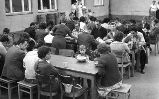 Drei lange Tische stehen hintereinander in einem Hof. An ihnen sitzen essende Frauen, Männer und Kinder.