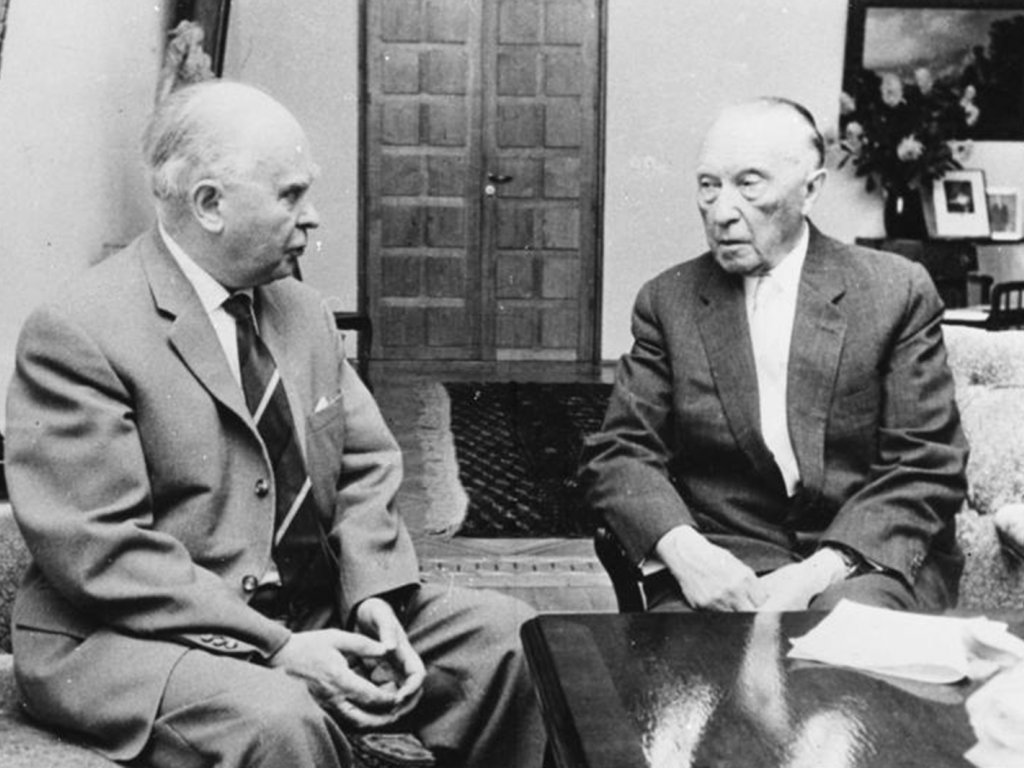 Kroll und Adenauer sitzen auf Sesseln an einem niedrigen Tisch und unterhalten sich mit ernster Miene.