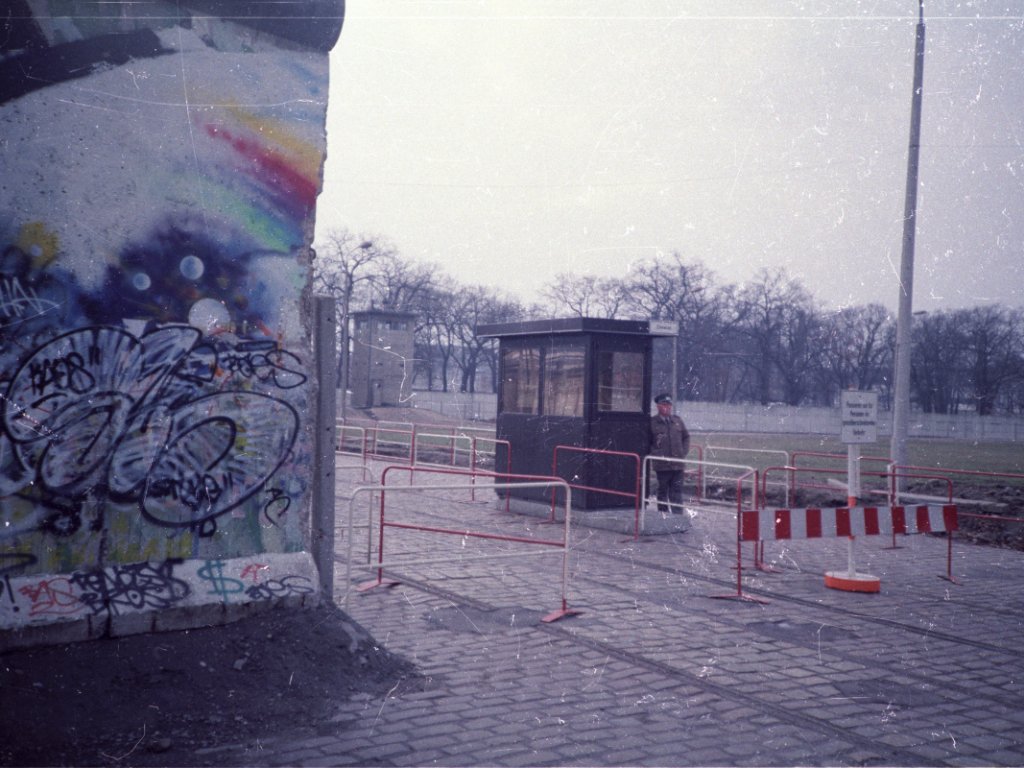 Ein Grenzsoldat steht vor seinem Kontrollhaus am Grenzübergang am Schlesischen Busch nahe dem Treptower Park. Die Grenzmauer ist mit Graffiti besprüht. Im Vordergrund sind Hinweisschilder erkennbar. Im Grenzstreifen steht eine Führungsstelle der Grenztruppen.