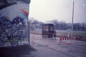 Ein Grenzsoldat steht vor seinem Kontrollhaus am Grenzübergang am Schlesischen Busch nahe dem Treptower Park. Die Grenzmauer ist mit Graffiti besprüht. Im Vordergrund sind Hinweisschilder erkennbar. Im Grenzstreifen steht eine Führungsstelle der Grenztruppen.