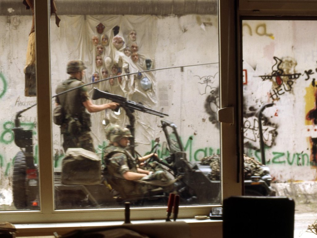 Soldaten fahren in einem offenen Jeep vorbei, das Bild ist aus dem Inneren der Galerie aufgenommen. Ein Soldat steht und hat die Hand am Abzug eines Gewehres. Im Hintergrund auf der Mauer sind installierte Gipsköpfe zu sehen.