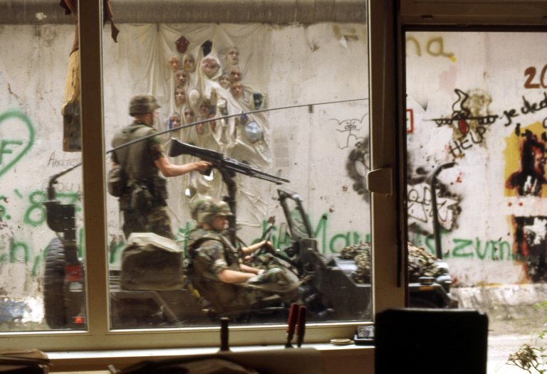 Soldaten fahren in einem offenen Jeep vorbei, das Bild ist aus dem Inneren der Galerie aufgenommen. Ein Soldat steht und hat die Hand am Abzug eines Gewehres. Im Hintergrund auf der Mauer sind installierte Gipsköpfe zu sehen.