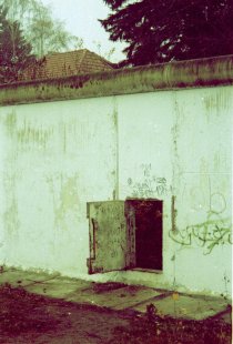 Ein Teil der Berliner Mauer ist groß im Bild. Im unteren Drittel eines Mauersegmentes befindet sich eine kleine Tür, die offen steht.