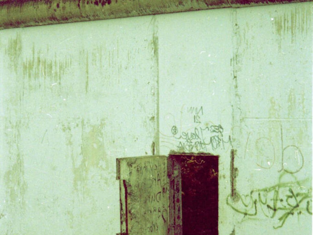 Ein Teil der Berliner Mauer ist groß im Bild. Im unteren Drittel eines Mauersegmentes befindet sich eine kleine Tür, die offen steht.
