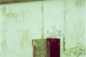 Ein Teil der Berliner Mauer ist gro&szlig; im Bild. Im unteren Drittel eines Mauersegmentes befindet sich eine kleine T&uuml;r, die offen steht.