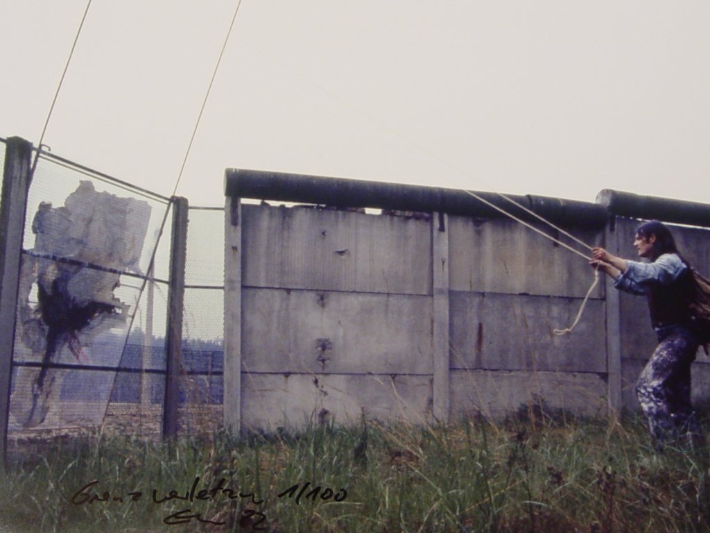 Links im Bild ist Elsners Gemälde zu sehen, das auf der anderen Seite des Streckmetallzaunes steht. Links im Bild steht Elsner und zieht an zwei Seilen, die an dem Gemälde befestigt sind. Die Grenze ist hier zur Hälfte mit dem Streckmetallzaun und zur Hälfte mit einer Betonmauer gesichert.