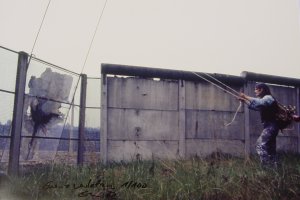 Links im Bild ist Elsners Gem&auml;lde zu sehen, das auf der anderen Seite des Streckmetallzaunes steht. Links im Bild steht Elsner und zieht an zwei Seilen, die an dem Gem&auml;lde befestigt sind. Die Grenze ist hier zur H&auml;lfte mit dem Streckmetallzaun und zur H&auml;lfte mit einer Betonmauer gesichert.