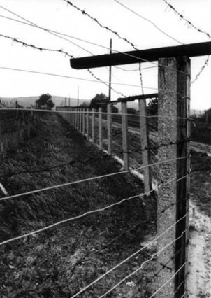 Nahaufnahme eines Zauns mit Stützpfeilern, Draht und Stacheldraht, der sich durch die hügelige Landschaft zieht.