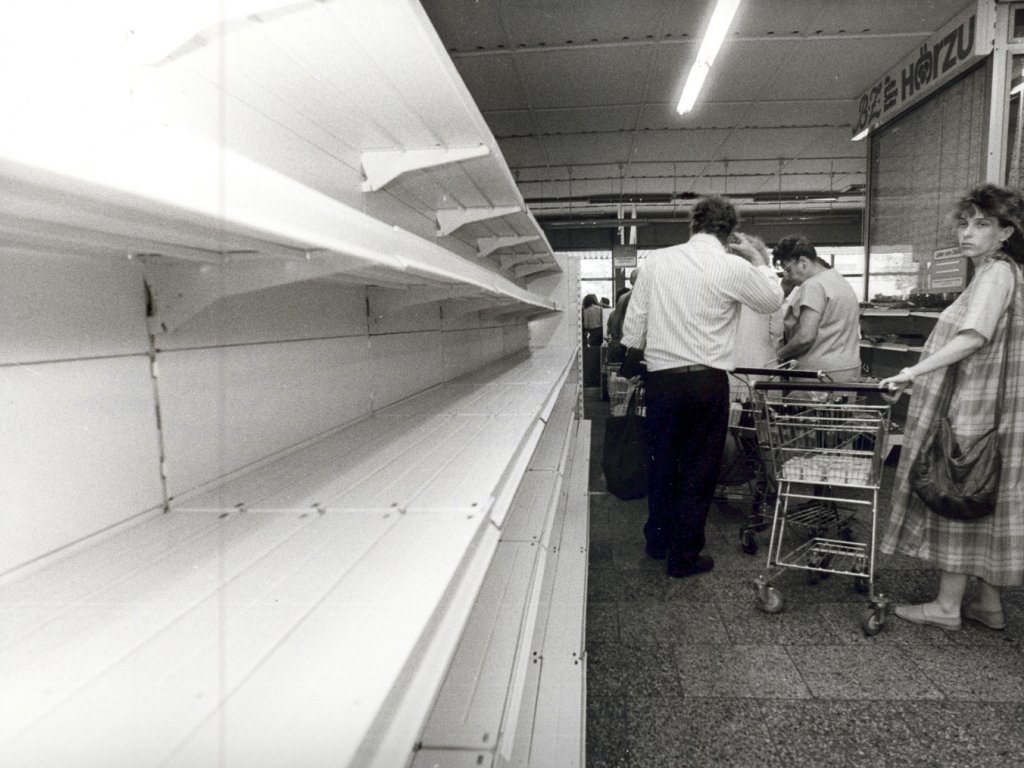 Wenige Tage vor der Währungsunion sind die Regale in den DDR-Kaufhallen leergefegt, 28. Juni 1990. Die linke Bildhälfte ist von einem leeren Kaufhallenregal eingenommen. Rechts davon steht eine Frau als letzte in der Kassenschlange. Außer einer Packung Toilettenpapier ist ihr Einkaufswagen leer.