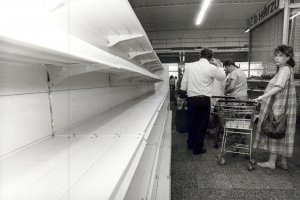 Die linke Bildhälfte ist von einem leeren Kaufhallenregal eingenommen. Rechts davon steht eine Frau als letzte in der Kassenschlange. Außer einer Packung Toilettenpapier ist ihr Einkaufswagen leer.
