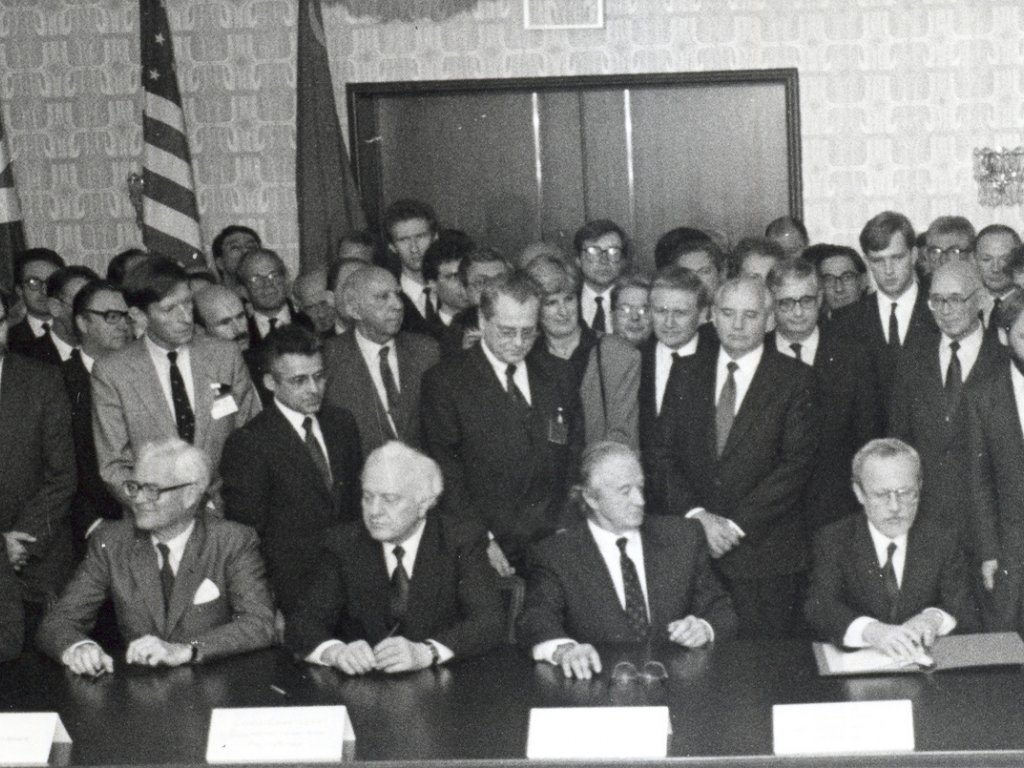 Die Außenminister sitzen am Tisch, vor Lothar de Maizière liegt der Vertrag. Hinter ihnen stehen dichtgedrängt Menschen, darunter Michail Gorbatschow. Sie blicken auf die Sitzenden.