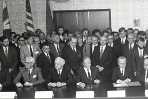 Die Außenminister sitzen am Tisch, vor Lothar de Maizière liegt der Vertrag. Hinter ihnen stehen dichtgedrängt Menschen, darunter Michail Gorbatschow. Sie blicken auf die Sitzenden.