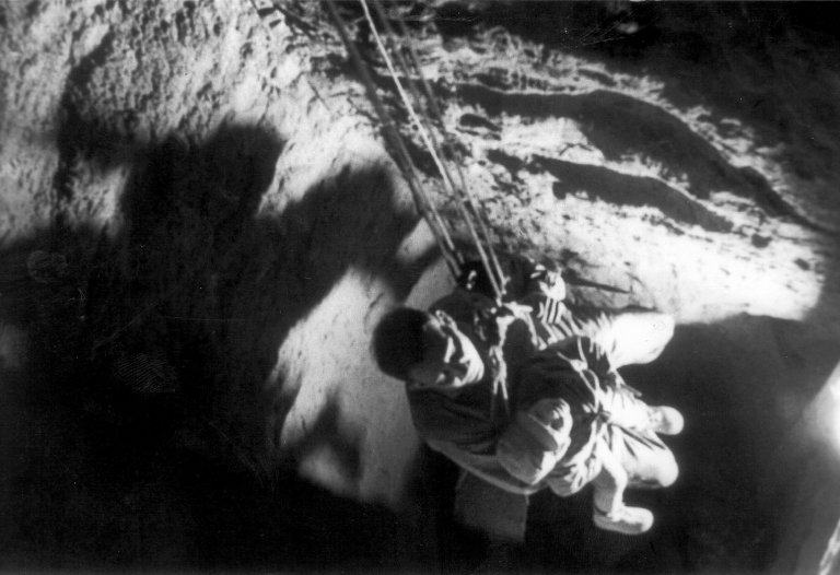 Der Blick geht von oben auf einen Mann mit einem Kind auf dem Schoß. Sie sitzen auf einem Brett und werden aus einem dunklen Schacht an Seilen in die Höhe gezogen.