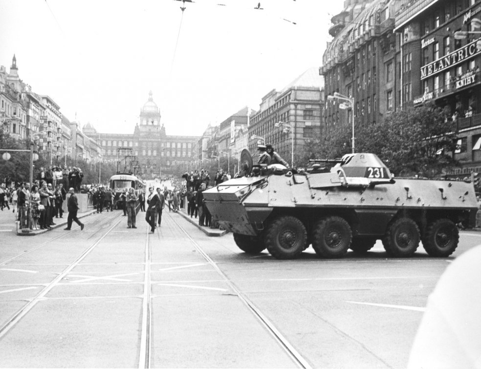 Im Vordergrund fährt ein Panzerwagen über die Straße. Er trägt eine Markierung mit der Zahl 231. Die Straße ist voller Menschen, die in Richtung des Militärfahrzeugs blicken. Im Hintergrund sind der Prager Wenzelsplatz und das Nationalmuseum zu erkennen.