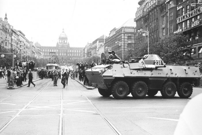 Im Vordergrund fährt ein Panzerwagen über die Straße. Er trägt eine Markierung mit der Zahl 231. Die Straße ist voller Menschen, die in Richtung des Militärfahrzeugs blicken. Im Hintergrund sind der Prager Wenzelsplatz und das Nationalmuseum zu erkennen.