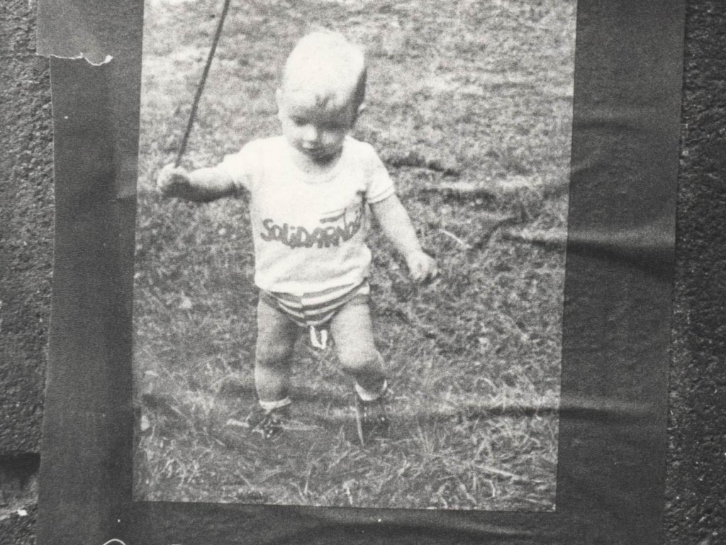 An einer Wand klebt ein Solidarnosc-Flyer. Darauf ist ein Foto von einem Kleinkind abgebildet. Es trägt ein T-Shirt, das mit dem Solidarnosc-Logo bedruckt ist. Unter dem Foto steht der Aufruf zum Kongress in polnischer Sprache.