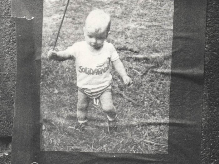 An einer Wand klebt ein Solidarnosc-Flyer. Darauf ist ein Foto von einem Kleinkind abgebildet. Es trägt ein T-Shirt, das mit dem Solidarnosc-Logo bedruckt ist. Unter dem Foto steht der Aufruf zum Kongress in polnischer Sprache.