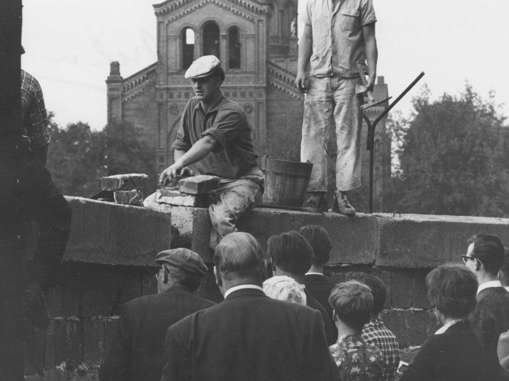 Ein Mann sitzt auf der Mauer und hantiert mit Ziegelsteinen. Hinter ihm steht ein zweiter Arbeiter mit einem Hammer in der Hand. Einige Menschen im Vordergrund beobachten die Arbeiten. Im Hintergrund ist ein Teil der St. Michael-Kirche erkennbar.