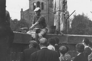 Ein Mann sitzt auf der Mauer und hantiert mit Ziegelsteinen. Hinter ihm steht ein zweiter Arbeiter mit einem Hammer in der Hand. Einige Menschen im Vordergrund beobachten die Arbeiten. Im Hintergrund ist ein Teil der St. Michael-Kirche erkennbar.