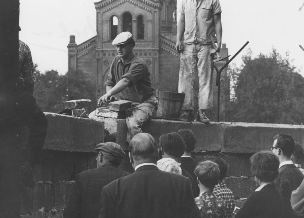 Ein Mann sitzt auf der Mauer und hantiert mit Ziegelsteinen. Hinter ihm steht ein zweiter Arbeiter mit einem Hammer in der Hand. Einige Menschen im Vordergrund beobachten die Arbeiten. Im Hintergrund ist ein Teil der St. Michael-Kirche erkennbar.