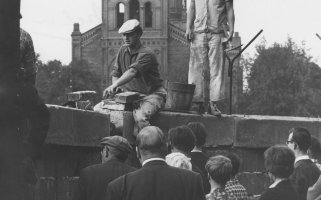 Ein Mann sitzt auf der Mauer und hantiert mit Ziegelsteinen. Hinter ihm steht ein zweiter Arbeiter mit einem Hammer in der Hand. Einige Menschen im Vordergrund beobachten die Arbeiten. Im Hintergrund ist ein Teil der St. Michael-Kirche erkennbar.